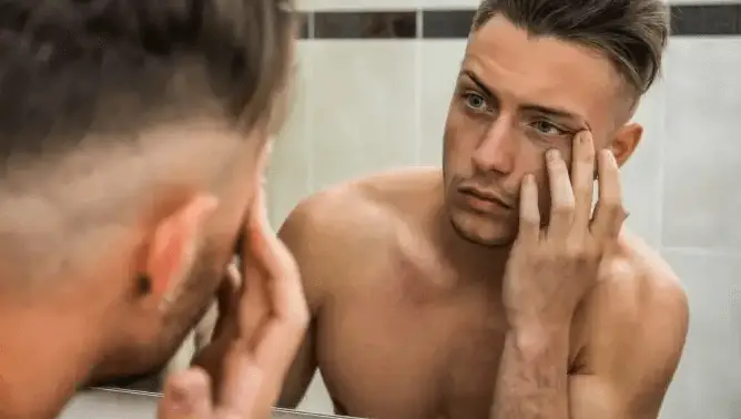 man looking in mirror at dark spots on his face.  showing need for a good men's facial product